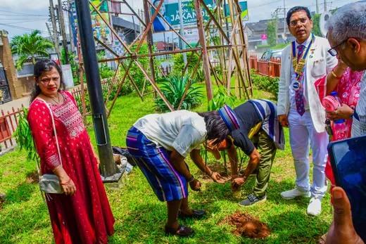 A member bent to the grass verge planting a sapling while the club looks on.